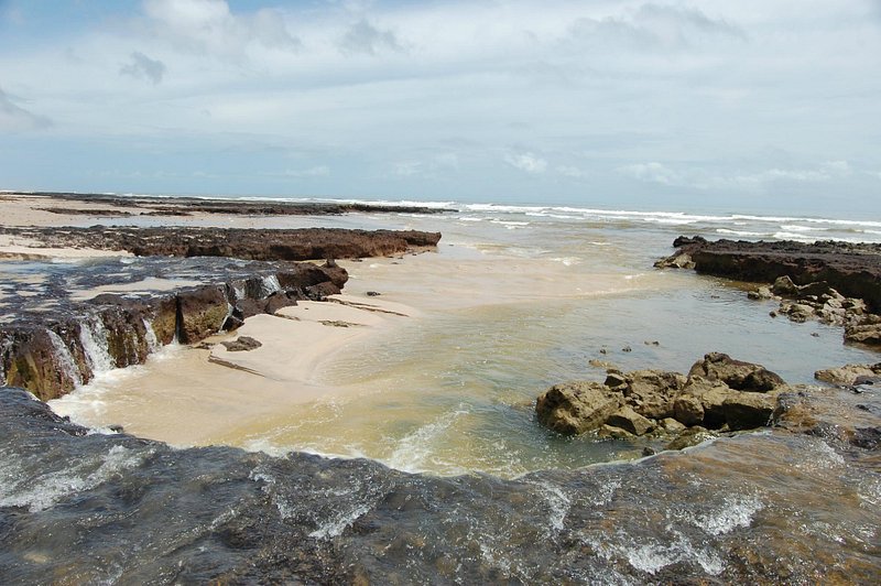 Poça natural entre rochas escuras na areia, ondas e céu nublado em Atins