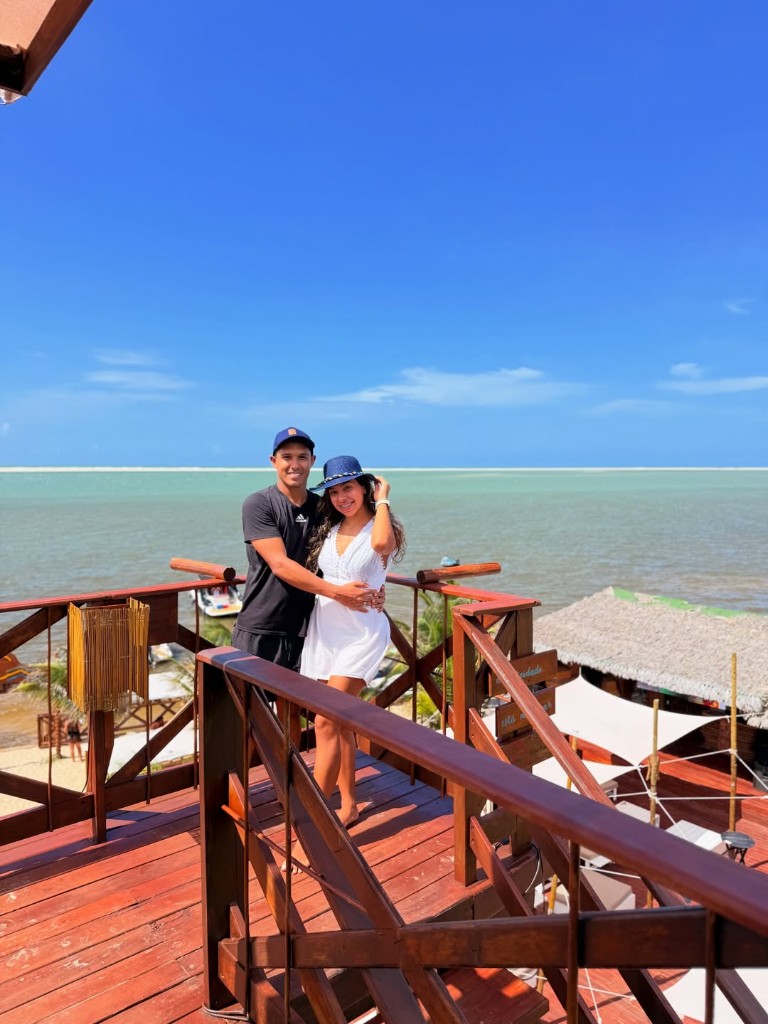 Casal no mirante de madeira com praia e mar azul ao fundo