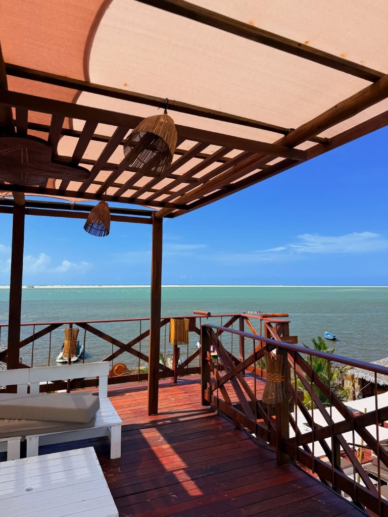 Deck com pérgola e vista para água turquesa, barcos e faixa de areia ao longe