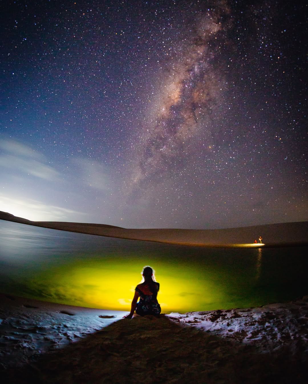 Visitante sentada na duna com lagoa iluminada e via láctea ao fundo